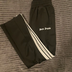  Palm Angels - Säljer ett par svarta byxor från Palm Angels med vita ränder längs sidorna. De har en elastisk midja och en avslappnad still. Byxorna är i mycket bra skick och passar både till vardags och ute. 