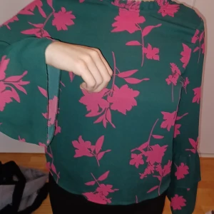 Grön blommig blus - Säljer en snygg grön blus med stora rosa blommor. Blusen har långa ärmar och en lös passform, perfekt för både vardag och fest. Materialet känns mjukt och bekvämt mot huden. Passar bra till både jeans och kjol!