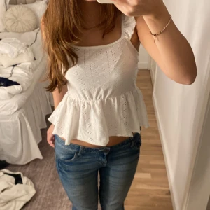 Vit ärmlös topp med volang - Säljer en superfin vit topp med volangdetaljer och öppen rygg. Den är ärmlös och har en söt knytning i ryggen. Perfekt för sommardagar eller en festlig kväll. Toppen är i ett lätt och luftigt material som känns skönt mot huden.