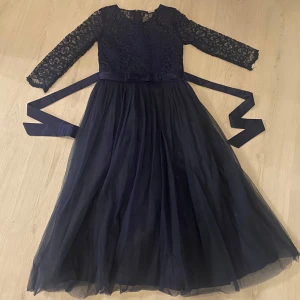 Mörkblå spetsklänning med lång kjol - Elegant mörkblå klänning med spetsdetaljer och lång kjol. Den har trekvartsärmar och ett vackert knytband i midjan. Perfekt för festliga tillfällen.