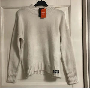 Vit stickad tröja från Superdry - Säljer en snygg vit stickad tröja från Superdry i storlek M. Den har en hög krage och ribbade muddar vid ärmslut och nederkant. Perfekt för kyliga dagar och passar till allt! 🧶✨