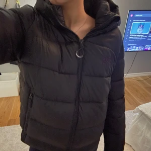 Svart dunjacka från Superdry - Säljer en svart dunjacka från Superdry i mycket bra skick. Jackan har en dragkedja framtill och två fickor med dragkedjor. Storleken är 44 alltså XL men den sitter precis som en S/M