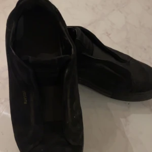 Svarta sneakers i mocka - Arigato Säljer ett par svarta sneakers i mocka. De är i bra skick och perfekta för vardagsbruk. Skorna har en stilren design utan snörning, vilket gör dem enkla att ta på och av. Passar till både jeans och mer uppklädda outfits.