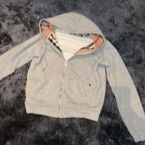 Grå huvtröja från Burberry - Säljer en snygg grå huvtröja från Burberry i storlek M. Den har en klassisk rutig insida i huvan och dragkedja framtill. Perfekt för höst och vår, med en stilren look som funkar till det mesta. Passar både till vardags och lite finare tillfällen.