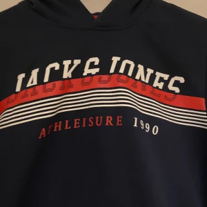 Mörkblå hoodie från Jack & Jones - Säljer en snygg mörkblå hoodie från Jack & Jones med tryck i vitt och rött. Perfekt för en avslappnad stil, med långa ärmar och en bekväm huva. Passar bra till både vardag och träning. Hör av dig vid intresse!