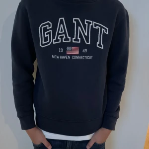 Mörkblå sweatshirt från GANT Strolek 146-152 - Säljer en snygg mörkblå sweatshirt från GANT med tryck 'New Haven Connecticut' och en liten amerikansk flagga. Tröjan har långa ärmar och är i mycket bra skick. Perfekt för en avslappnad stil!