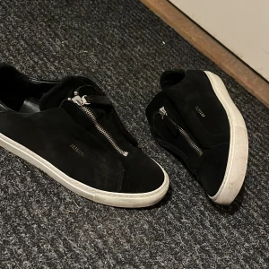 Svarta Axel argatos med dragkedja. - Säljer ett par svarta sneakers från Axel Arigato i bra skick. De är gjorda i mocka och har en snygg dragkedja på skorna. 