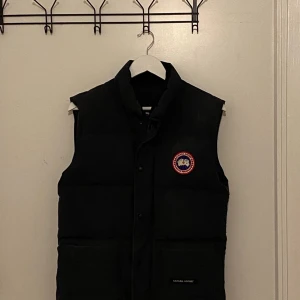 Svart dunväst från Canada Goose - Säljer en svart dunväst från Canada Goose i mycket bra skick. Västen har en klassisk design och stängs med dragkedja och knappar. Perfekt för kyliga höst- och vinterdagar. Passar både till vardags och mer uppklädda tillfällen.