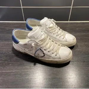 Säljer ett par vita sneakers från Philippe Model i bra skick. De har blå detaljer på hälen och klassisk snörning. Skorna kommer med originalförpackning och påse. Storleken är 40-41