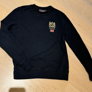 Svart tröja från Morris strl XS - Stilren svart collegetröja från Morris med ett broderat emblem på bröstet. Tröjan är i mycket fint skick, knappt använd. Nypriset var 1299kr