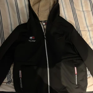Svart jacka med teddyfoder - Säljer en svart jacka från Hilfiger i storlek M. Den har ett mysigt teddyfoder som håller dig varm under kalla dagar. Jackan har dragkedja och två praktiska fickor framtill. Perfekt för höst och vinter!