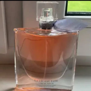 La Vie Est Belle Eau de Parfum - Säljer en elegant flaska av La Vie Est Belle Eau de Parfum från Lancôme. Flaskan är genomskinlig med en vacker rosa vätska inuti och har en stilren fyrkantig kork. Den har en liten dekorativ rosett vid halsen som ger en extra touch av lyx. Perfekt för den som vill dofta fantastiskt varje dag! 🌸