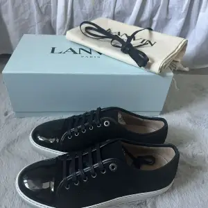 Säljer ett par snygga svarta sneakers från Lanvin i nytt skick. De har en glansig tå och är gjorda i skinn med snörning. Perfekta för både vardag och fest! Kommer med originalkartong och dustbag. Perfekt julklapp.