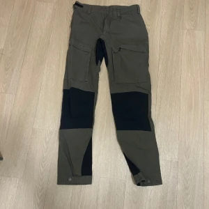 Gröna byxor från Lundhags - Säljer ett par gröna byxor från Lundhags i bra skick. De har en praktisk design med flera fickor och förstärkningar i svart på knän och insida av benen. Perfekta för utomhusaktiviteter och friluftsliv. Byxorna har knappar vid bensluten för justering.