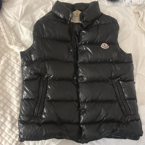 Svart dunväst från Moncler - Säljer en snygg svart dunväst från Moncler i mycket bra skick. Västen har en glansig yta med dragkedja och knappar framtill. Perfekt för höst och vinter, och den har en klassisk Moncler-logga på bröstet. Västen är både stilren och varm, storlek 1 som motsvarar S💗💗💗