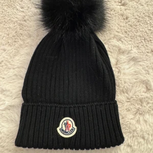 Äkta Moncler mössa  - Snygg svart stickad mössa från Moncler med en stor fluffig tofs på toppen. Den har en broderad Moncler-logga framtill och är perfekt för kalla vinterdagar. Mössan är i mycket bra skick och passar perfekt för både vardag och andra tillfällen. Väldigt liten i storlek 