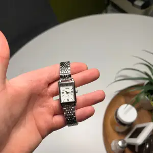 Säljer en stilren och elegant klocka från Axcent i silverfärg. Den har en rektangulär urtavla med svarta visare och markeringar. Armbandet är i metall med en klassisk länkdesign. Perfekt för både vardag och fest!