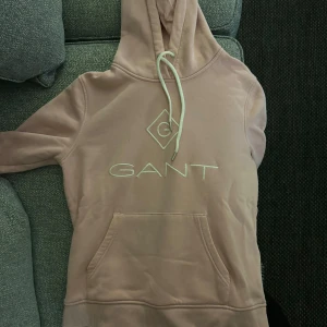 Rosa hoodie från GANT - Säljer en snygg beige hoodie från GANT i bra skick. Den har en stor ficka fram och en justerbar huva med dragsko. Perfekt för en avslappnad stil och passar till alla årstider. GANT-loggan är broderad på framsidan. Superbekväm och stilren!