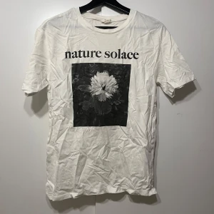 Vit t-shirt med tryck - Säljer en vit t-shirt med texten 'nature solace' och ett svartvitt blommotiv på framsidan. Perfekt för en avslappnad stil och passar bra till jeans eller shorts. T-shirten är i bomull och har en normal passform. Perfekt för sommardagar eller som ett lager under en jacka. Passar också S
