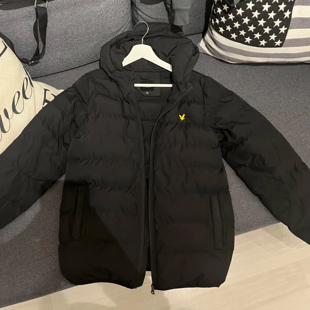 Säljer en svart pufferjacka från Lyle & Scott i mycket bra skick. Jackan har en praktisk huva och dragkedja framtill. Perfekt för höst och vinter, håller dig varm och stilren. Den har en liten gul logga på bröstet som ger en snygg detalj. Passar perfekt för både vardag och lite kyligare dagar. Jackan passar barn i ca 13 års ålder . Jackor.