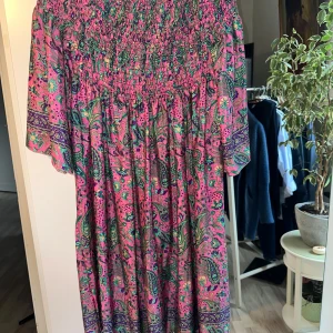 Färgglad klänning i polysilke - Säljer en superfin klänning i polysilke med ett färgglatt paisleymönster i rosa, lila och grönt. Klänningen har en smockad överdel och trekvartsärmar, perfekt för sommarens fester. Tillverkad i Indien och känns både luftig och bekväm. Passar perfekt för en bohemisk look!