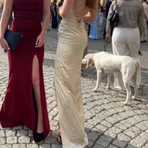 Champagnefärgad långklänning med öppen rygg - Säljer en elegant champagnefärgad långklänning i satin. Klänningen har en tight passform med en slits framtill och en vacker öppen rygg med snörning. Perfekt för festliga tillfällen som bal eller nyår. Den är axelbandslös och ger en lyxig känsla. Hör av dig vid intresse! Köpt för 999kr