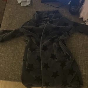 Mjuk morgonrock med stjärnor från KappAhl - Säljer en supermjuk och mysig morgonrock från KappAhl i storlek 134/140. Den är mörkgrå med svarta stjärnor och har ett praktiskt bälte i midjan. Perfekt för kalla morgnar eller mysiga kvällar hemma. 🖤Har inte bandet, den har försvunnit