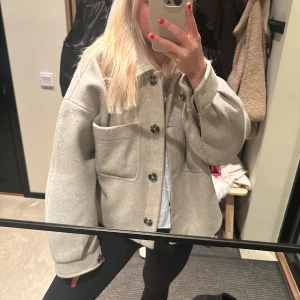 Beige overshirt med knappar - Säljer en snygg beige/grå kappa i mycket bra skick. Den har stora knappar framtill och två praktiska bröstfickor. Perfekt för höst och vår, och funkar både till vardags och lite mer uppklätt. Passformen är normal och den är långärmad. Supermysig och stilren!