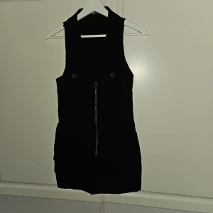 V-ringning svart klänning  - Nytt Svart klänning med guldfärgad dragkedja framtill och två fickor framtill Klänningen är utan ärmar och har en v-ringning . Hällor för att kunna ha skärp I Längd 78 cm