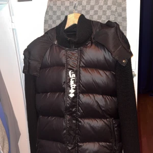 Svart pufferjacka från This is Sixties - Säljer en svart pufferjacka från This is Sixties i storlek S. Jackan har en cool design med stickade ärmar och en hög krage. Den är perfekt för höst och vinter med sin varma vaddering och dragkedja framtill. Passar både till vardags och lite mer uppklädda tillfällen.