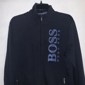 Mörkblå kofta Hugo Boss - Säljer en mörkblå kofta från Hugo Boss i storlek M. Jackan har en dragkedja framtill och ett stort BOSS-tryck i blått på bröstet. Den är i mycket bra skick har knappt använt den.