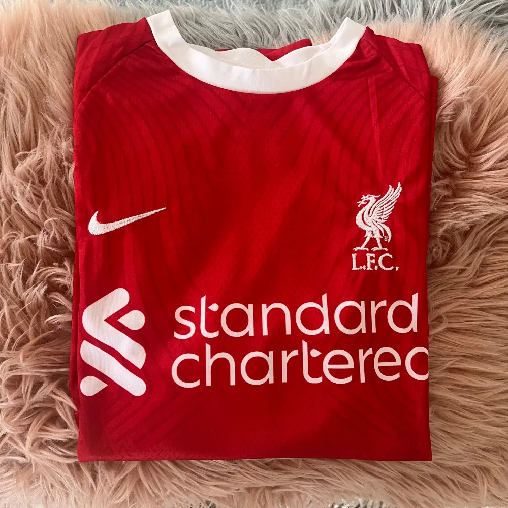 Säljer en röd Liverpool fotbollströja från Nike med vit krage och vita detaljer. Tröjan har korta ärmar och är i nyskick. Den har 'VIRGIL' och nummer 4 tryckt på baksidan. Perfekt för alla Liverpool-fans!. T-shirts.