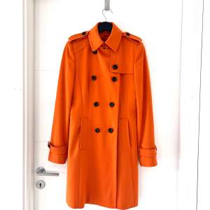 Säljer en snygg orange trenchcoat från Hugo Boss. Den har en klassisk dubbelknäppning med bruna melerade knappar och axelklaffar. Jackan är långärmad och har en stilren design med ett bälte baktill. Perfekt för höst och vår! 