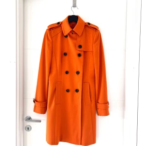 Orange trenchcoat från Hugo Boss - Säljer en snygg orange trenchcoat från Hugo Boss. Den har en klassisk dubbelknäppning med bruna melerade knappar och axelklaffar. Jackan är långärmad och har en stilren design med ett bälte baktill. Perfekt för höst och vår! 