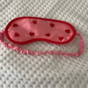 Rosa sovmask med hjärtan - Supergullig rosa sovmask med röda hjärtan. Den har en elastisk rem som är täckt med rosa tyg för extra komfort. Perfekt för en god natts sömn eller en mysig tupplur!