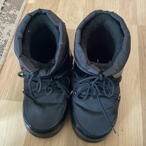 Svarta moon boots - Säljer ett par svarta moon boots i syntetmaterial. De är perfekta för vintern med sin varma och vadderade insida. Skorna har snörning framtill och en glansig yta som ger dem en cool look. Perfekta för kalla dagar!