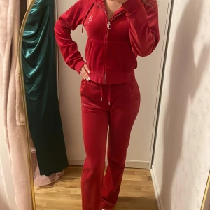 Röd velour hoodie från Juicy Couture - Säljer ett supermjukt och snyggt rött velour set från Juicy Couture. Den har en dragkedja framtill och är dekorerad med glittriga stenar som bildar texten 'Juicy' på baksidan av både tröjan och byxan. Perfekt för en avslappnad och stilren look. Passar perfekt till höst och vinter! är i väldigt bra skick. skriv för frågor!!
