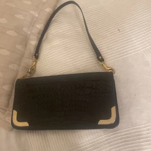 Svart axelväska med gulddetaljer - Säljer en snygg svart axelväska med krokodilmönster och stiliga gulddetaljer i hörnen. Väskan har en avtagbar axelrem och är perfekt för både vardag och fest. Den är i bra skick!!💕 typ aldrig använd!🌸 kontakta innan köp man får gärna buda