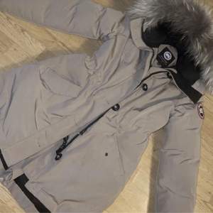 Säljer en supervarm och snygg beige vinterjacka från Canada Goose. Den har en fluffig pälskrage och långa ärmar, perfekt för kalla vinterdagar. Jackan har knappar för extra skydd mot kylan. Den är i nyskick och redo för nya äventyr! 