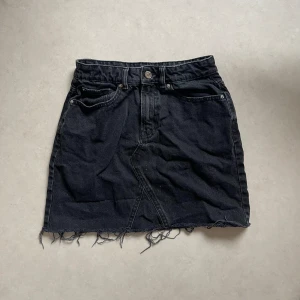 Svart jeanskjol med fransig kant - Säljer en cool svart jeanskjol med en fransig kant som ger en avslappnad look. Kjolen har en klassisk femficksdesign och stängs med en knapp och dragkedja framtill. Perfekt för en casual outfit eller en utekväll med vänner!