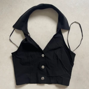 Svart korsettopp med knappar - Snygg svart korsettopp med tre knappar framtill. Den har en unik krage och smala axelband som ger en stilren look. Perfekt för festliga tillfällen!