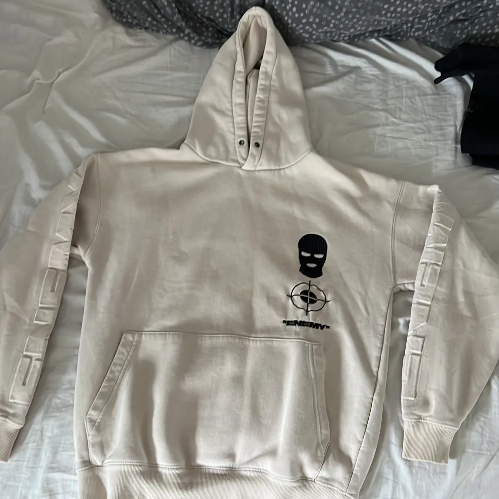 Säljer en cool beige hoodie från Black Squad i storlek XS. Den har en oversized passform och är perfekt för en avslappnad stil. På framsidan finns en tryckt design med en mask och texten 'ENEMY'. Baksidan har en stor bild av en hund med texten 'ENEMIES'. Hoodien har en stor ficka framtill och långa ärmar med ribbade muddar. Perfekt för höst och vinter!. Tröjor & Koftor.