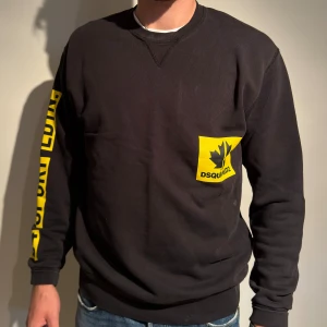 Svart sweatshirt från Dsquared2 - Säljer en svart sweatshirt från Dsquared2 med coola gula detaljer. Tröjan har en gul logga på bröstet och text längs ärmen. Perfekt för en sportig look eller bara för att chilla i. Den är långärmad och gjord i ett bekvämt material. Passar perfekt till jeans eller joggers!
