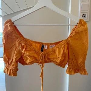 Orange croppad topp från H&M - Säljer en superfin orange croppad topp från H&M. Den har korta ärmar med volangdetaljer och en söt knytning framtill. Perfekt för sommaren och passar både till fest och vardag. Materialet är lätt och bekvämt.