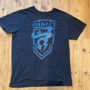 Svart t-shirt från Oakley - Säljer en svart t-shirt från Oakley med ett coolt tryck i blått och vitt på framsidan. T-shirten har en normal passform och är tillverkad i bomull, vilket gör den bekväm att bära. Perfekt för vardagsbruk eller träning! Storlek XL men sitter som M