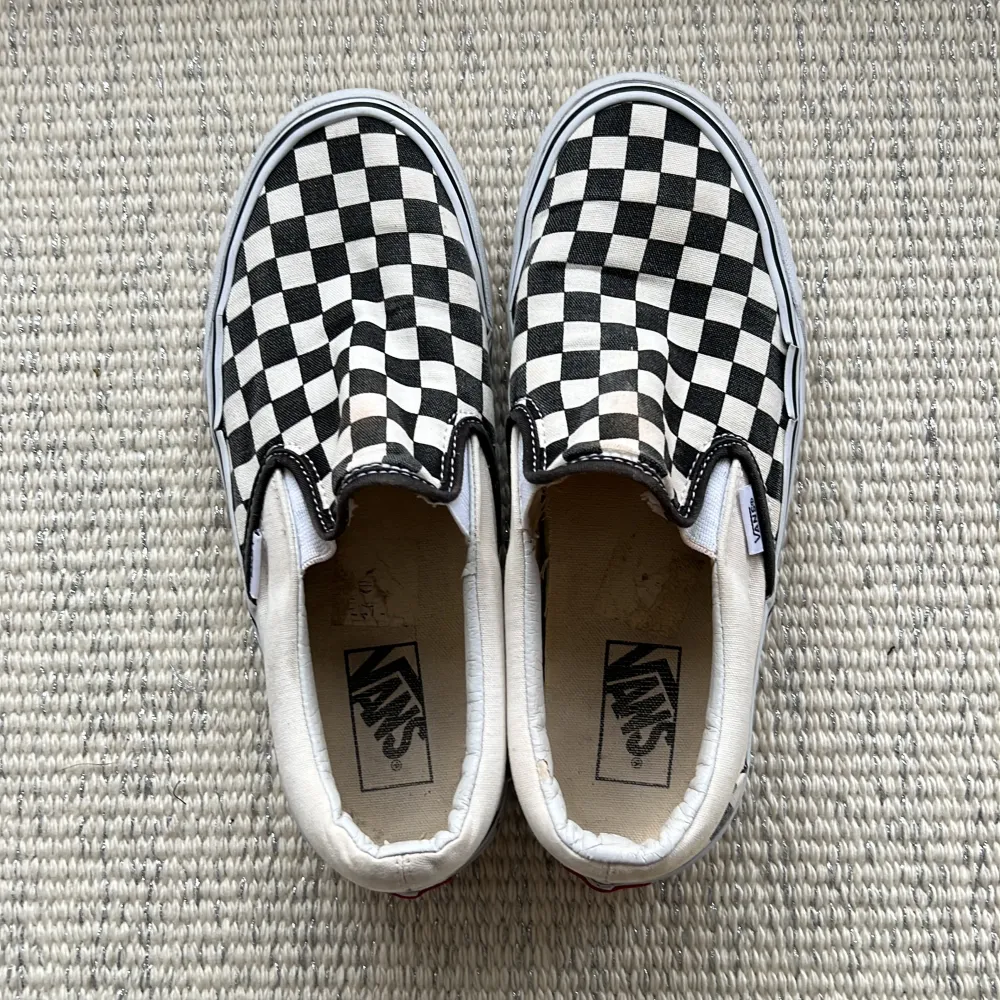 Säljer ett par klassiska svartvita slip-on sneakers från Vans med det ikoniska rutiga mönstret. Skorna är i bra skick och perfekta för en avslappnad stil. De har en bekväm passform och är lätta att ta på och av.. Skor.