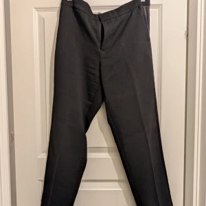 Svarta kostymbyxor - Snygga svarta kostymbyxor med en klassisk design. De har en rak passform och är perfekta för en stilren look. Byxorna har en dragkedja framtill och hällor för bälte. Storlek 38/40