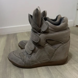 Isabel Marant liknande skor - Snygga grå sneakers med kilklack och kardborreband. Skorna har en unik design med hög ankel och flera remmar för en cool look. Perfekta för att ge lite extra höjd och stil till din outfit. Färgen är lite grå/brun.