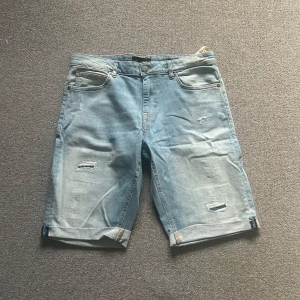 Ljusblå jeansshorts från New Yorker - Säljer ett par ljusblå jeansshorts från New Yorker med slitna detaljer och uppvikta benslut. Shortsen har en klassisk femficksdesign och stängs med knapp och dragkedja. Perfekta för en avslappnad stil.