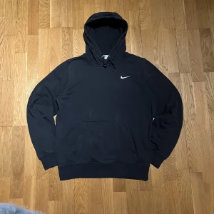 Svart hoodie från Nike - Säljer en klassisk svart hoodie från Nike med den ikoniska swoosh-loggan på bröstet. Tröjan har en stor ficka framtill och justerbar huva med dragsko. Perfekt för en avslappnad stil.